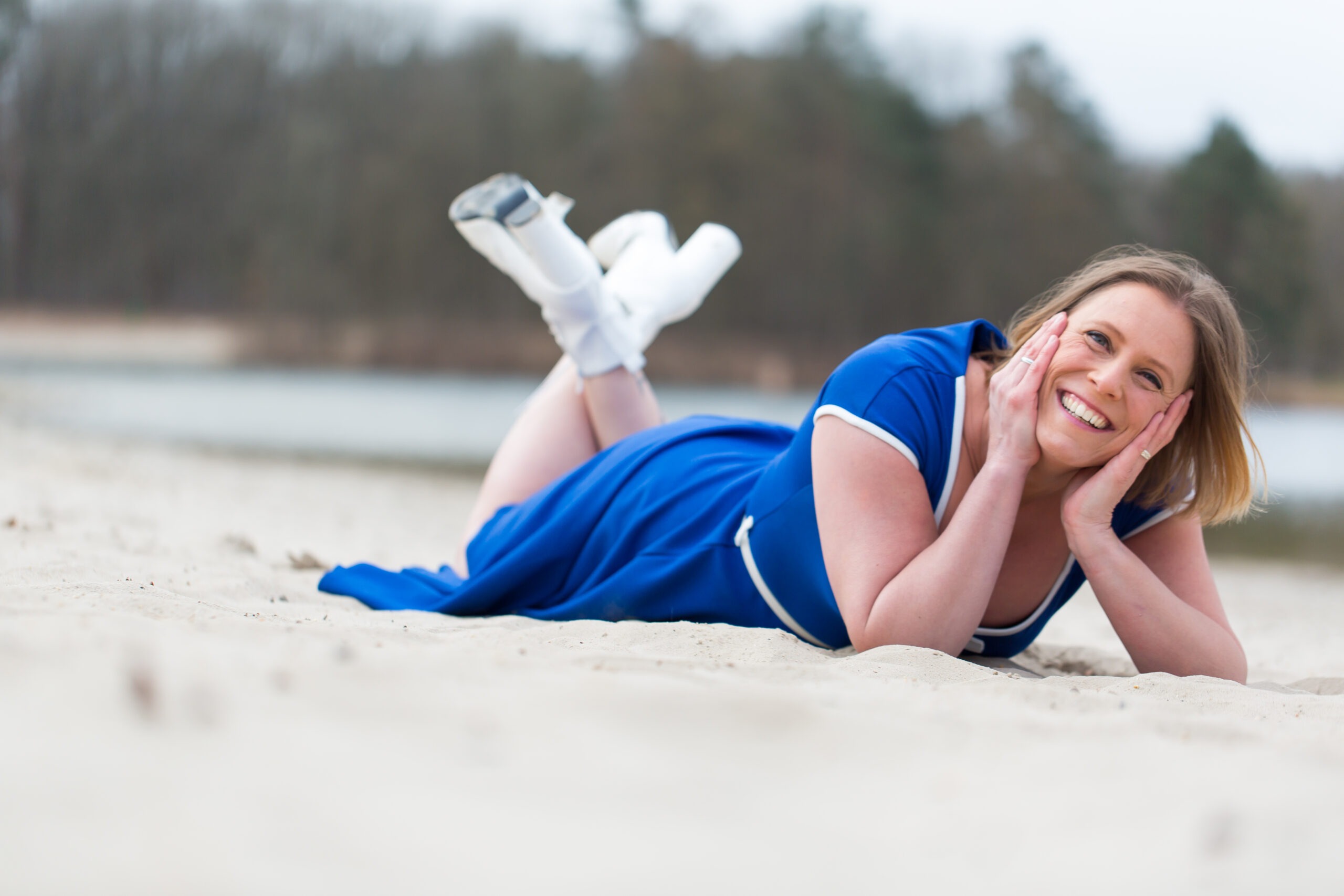 Op de foto zie je Claudia Stinne lachend op haar buik in het zand liggen. Ze heeft haar onderbenen gekruist omhoog en steunt met haar gezicht in haar handpalmen.