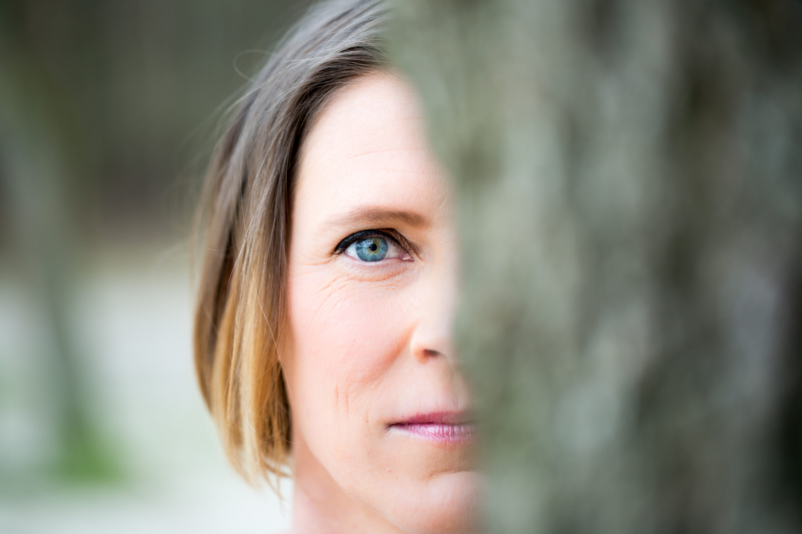 Claudia Stinne - auteur en coach Op de foto zie je de helft van het gezicht van Claudia Stinne. Ze kijkt je recht aan met een zeer blauwe iris. De rest van haar gezicht is buiten beeld, door een boom die out of focus is